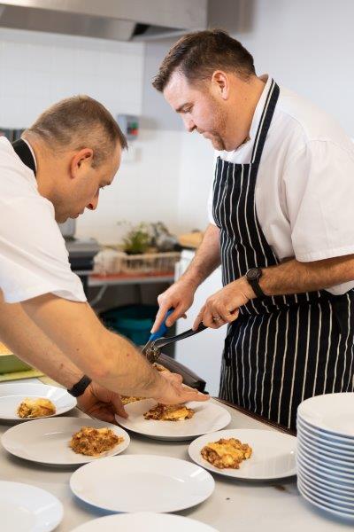two chefs in a commercial kitchen, serving several plates of food